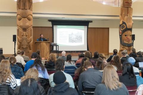 Group of people attending a workshop, with a presenter in the front in a podium and screen behind them